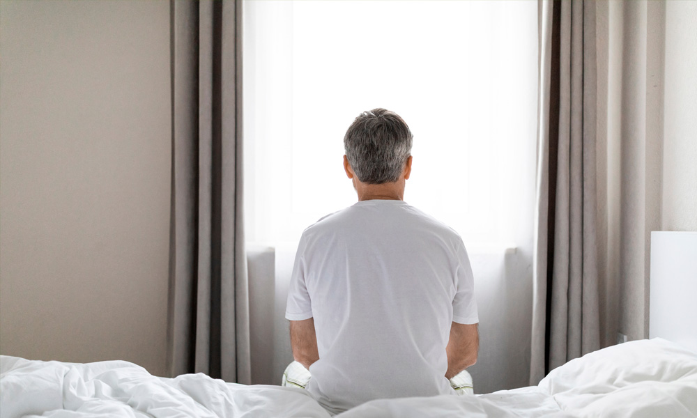 hombre sentado en la cama viendo hacia la vetana cerrada